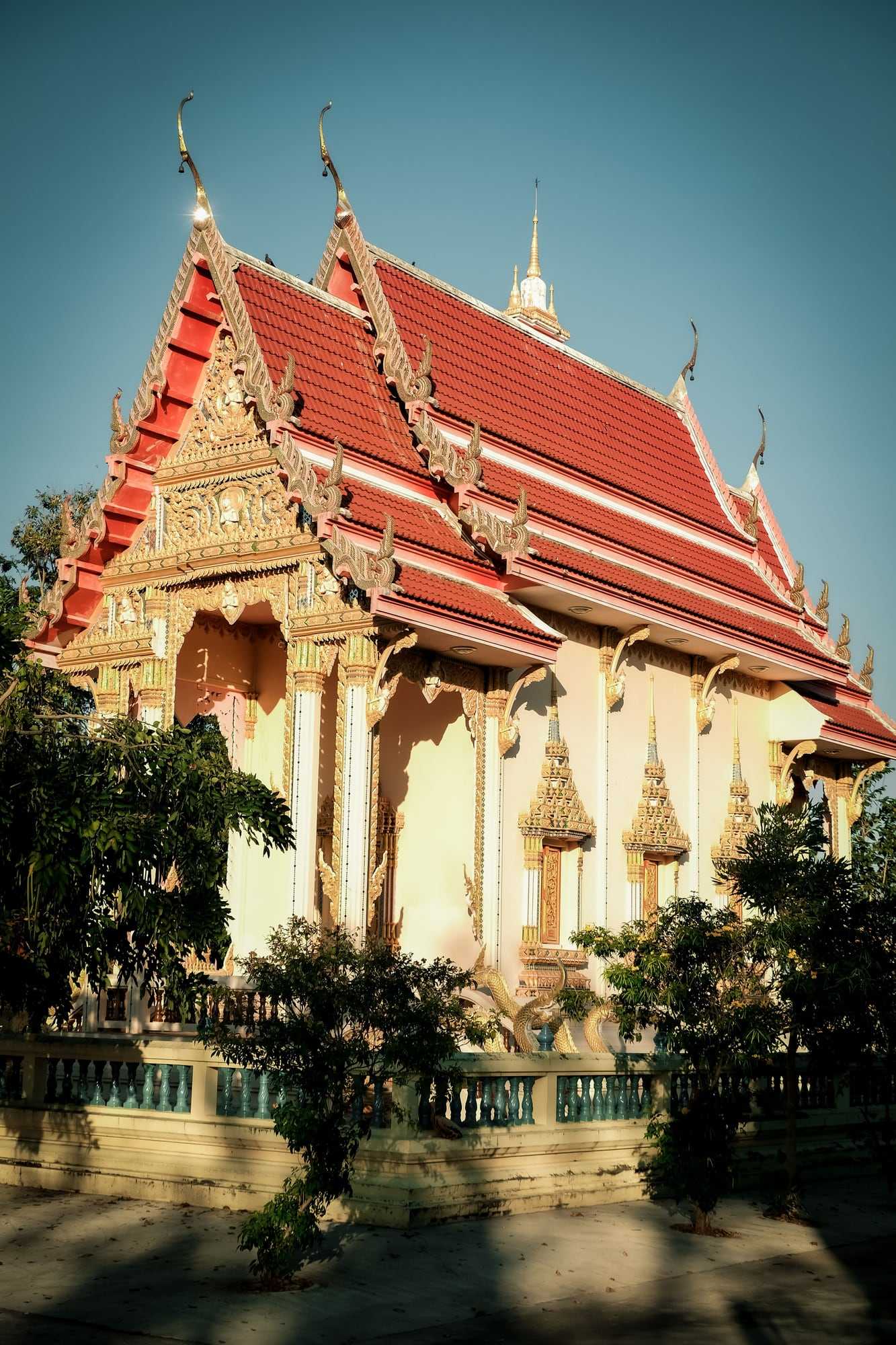 A Thai temple hall with a tiered red and gold roof, in warm afternoon light
